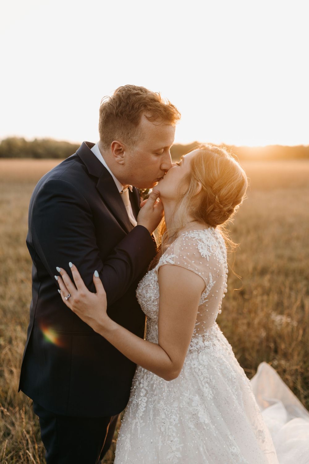 Bride and groom kissing passionately in sunlit wheat field during golden hour wedding photography session.