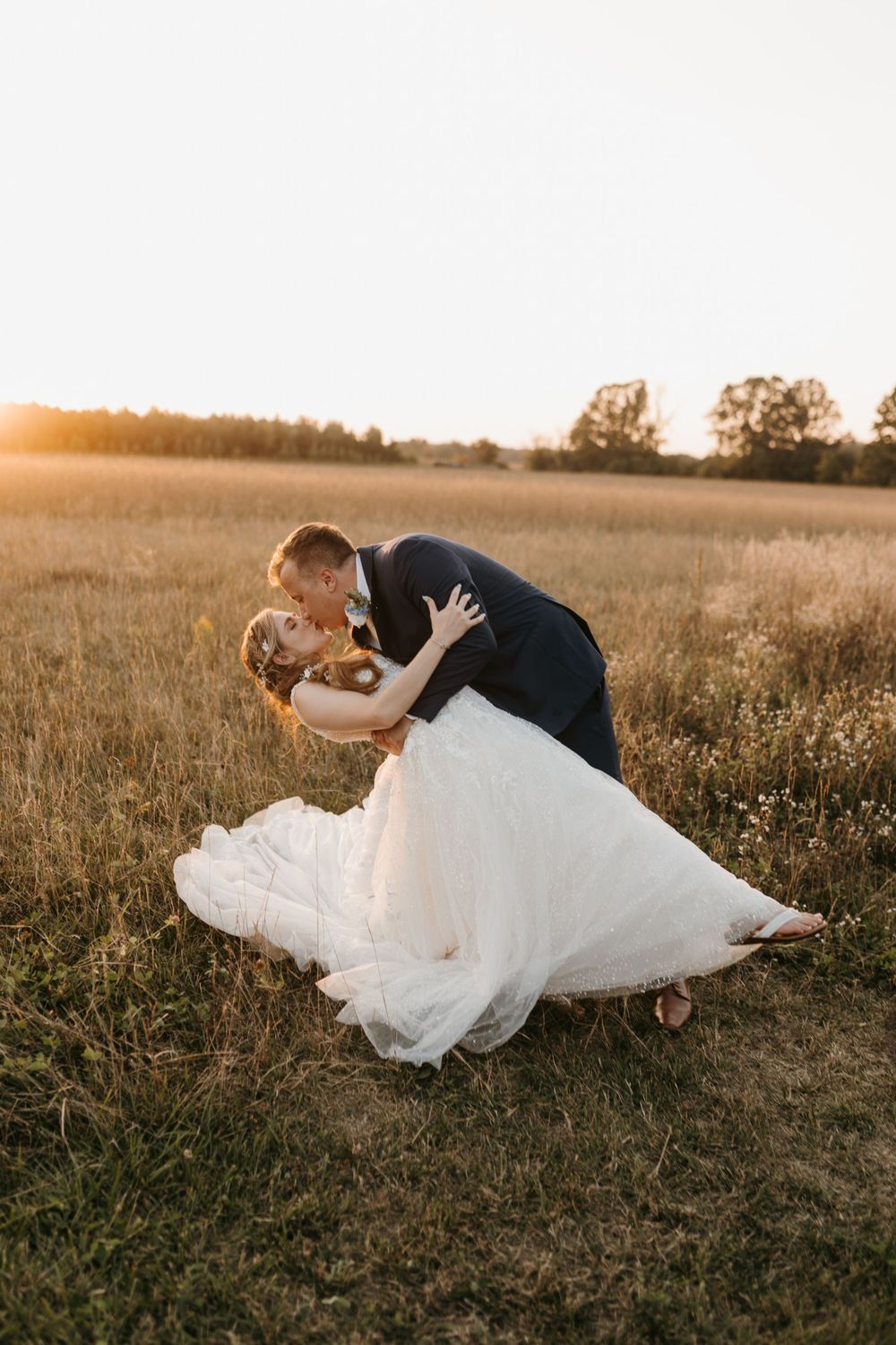 Groom dipping bride in flowing white gown for a romantic kiss in a sunlit field.