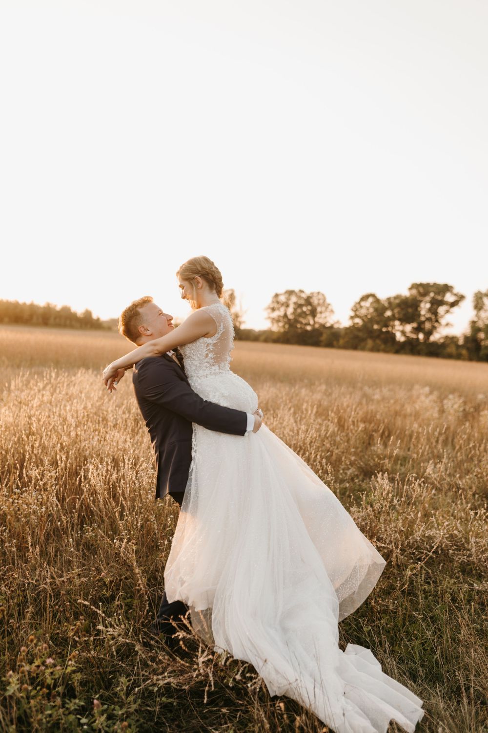 Groom lifting bride in white lace gown in sunlit wheat field during golden hour wedding shoot.
