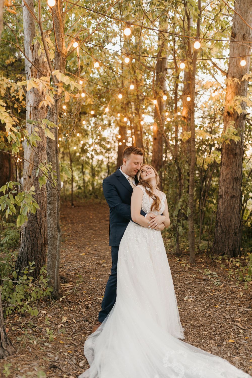 Bride and groom embracing under string lights in a beautiful autumn forest setting.