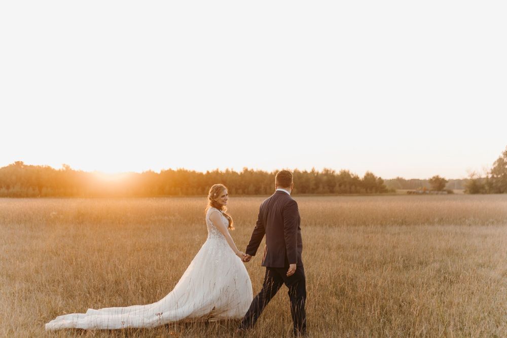 Bride and groom walking hand in hand through golden field during stunning sunset.