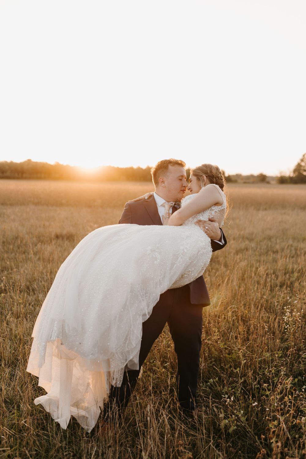 Groom carrying bride in white lace gown through golden sunlit field at sunset.