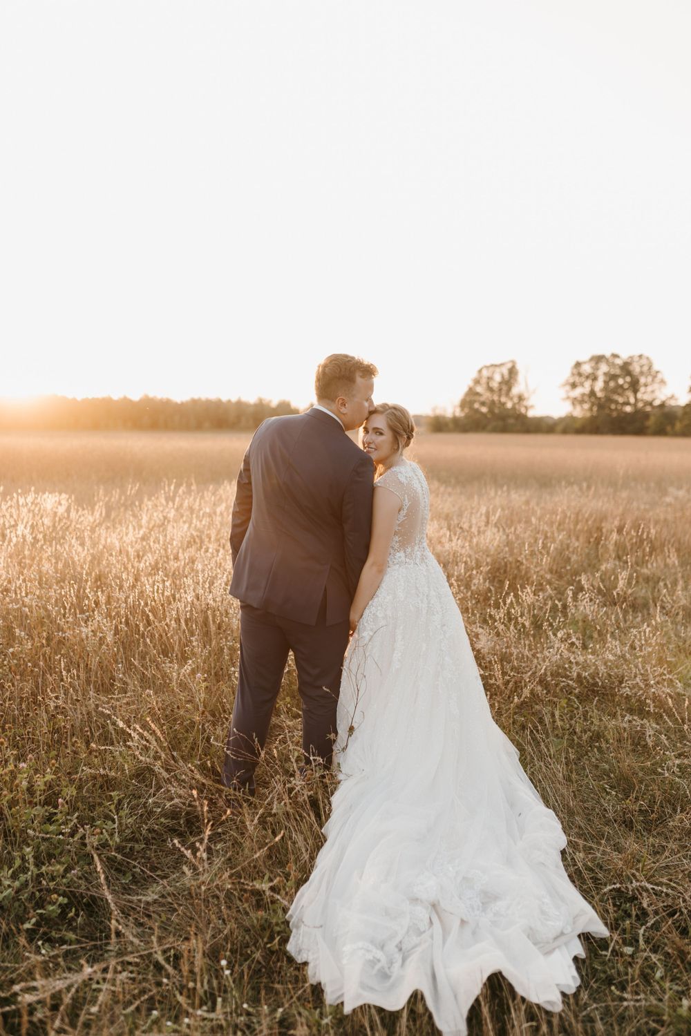Groom kissing bride on forehead in golden wheat field during romantic sunset wedding portrait.