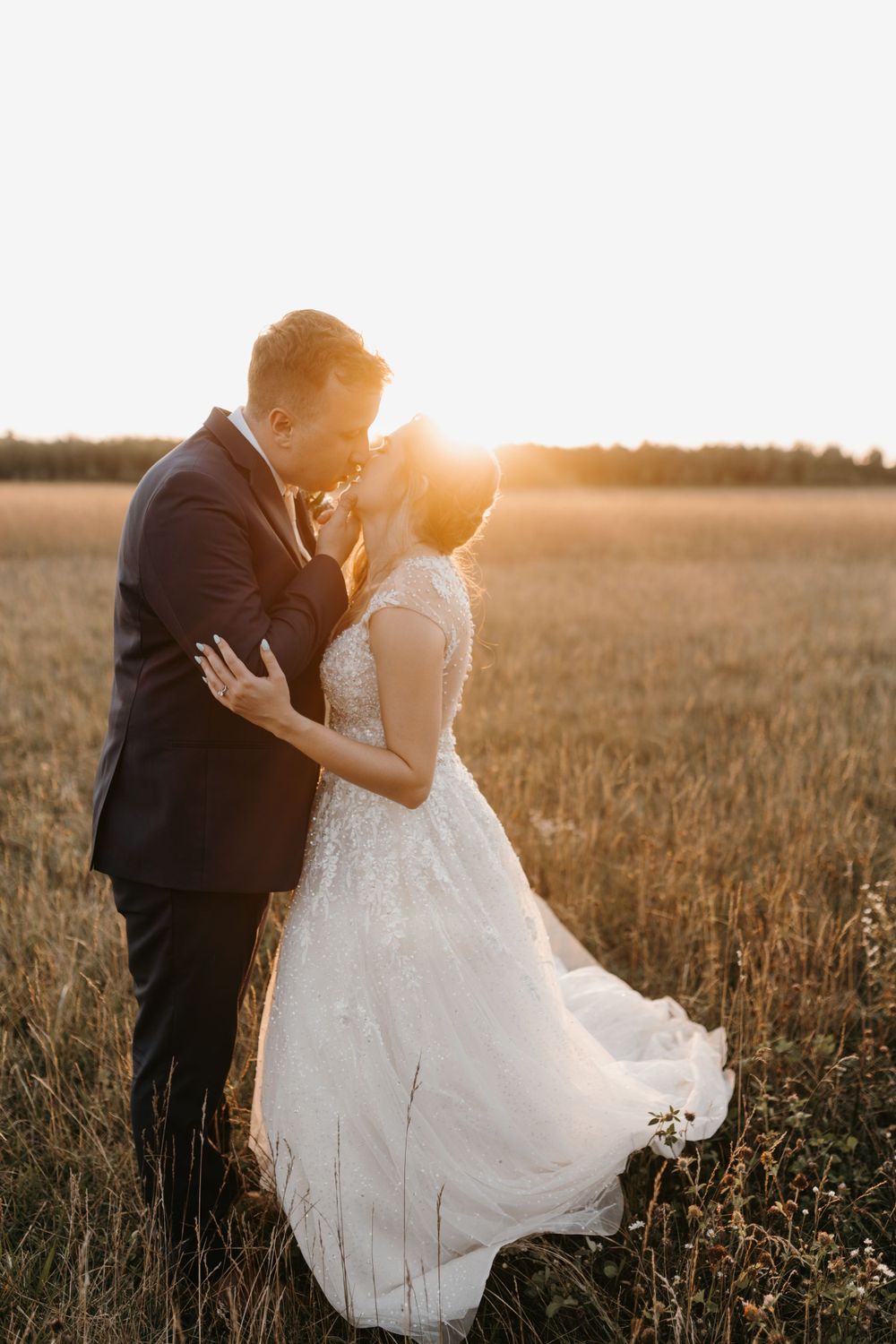 Newlyweds sharing tender kiss in golden wheat field with stunning warm sunset glowing behind them.