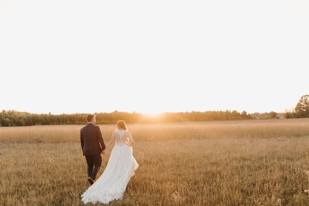 Bride and groom holding hands walking into stunning golden sunset across open wheat field.