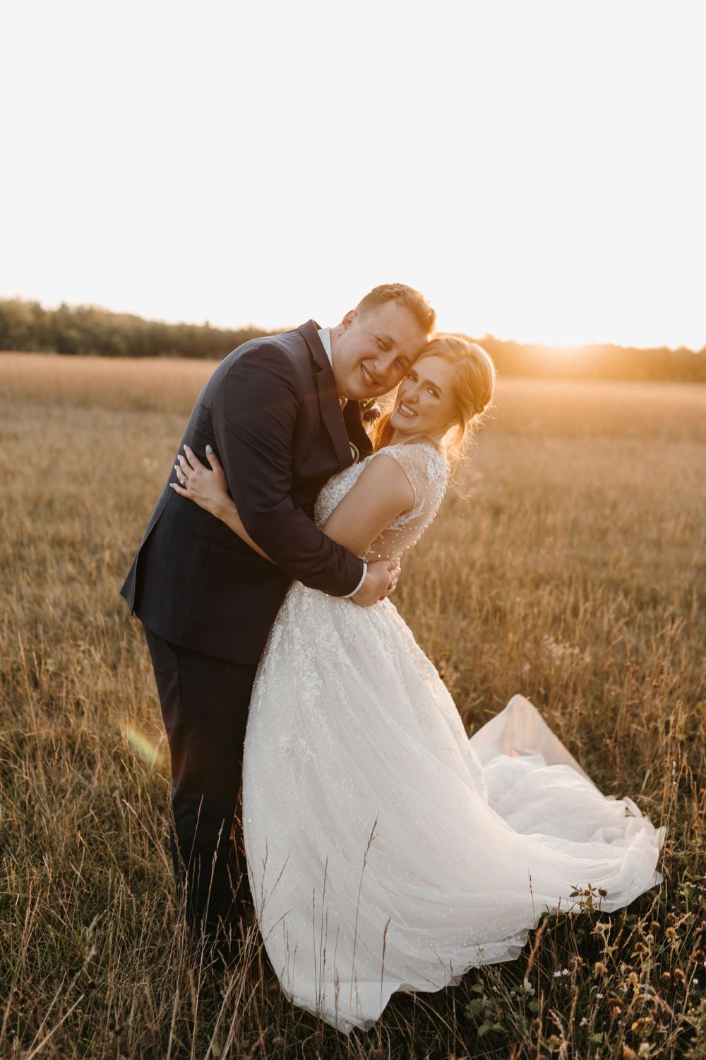 Bride and groom embracing in golden field at sunset, bride wearing lace wedding gown.