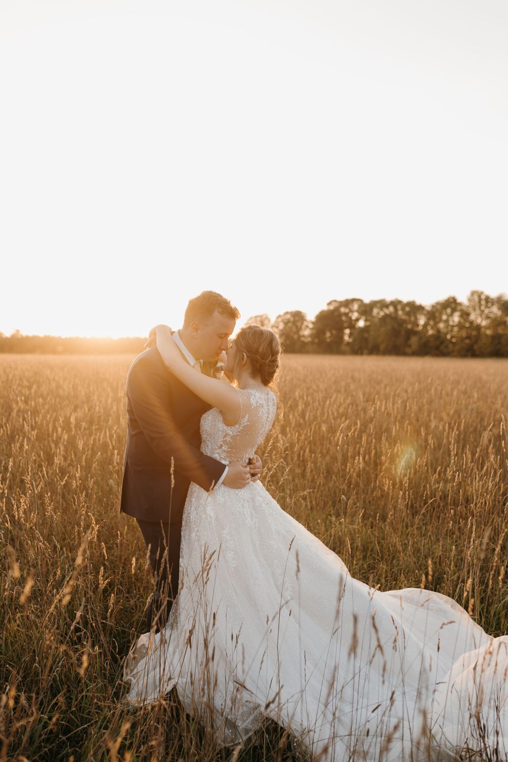 Newlyweds embrace romantically in a golden wheat field at sunset, brides lace gown and train spread beautifully.