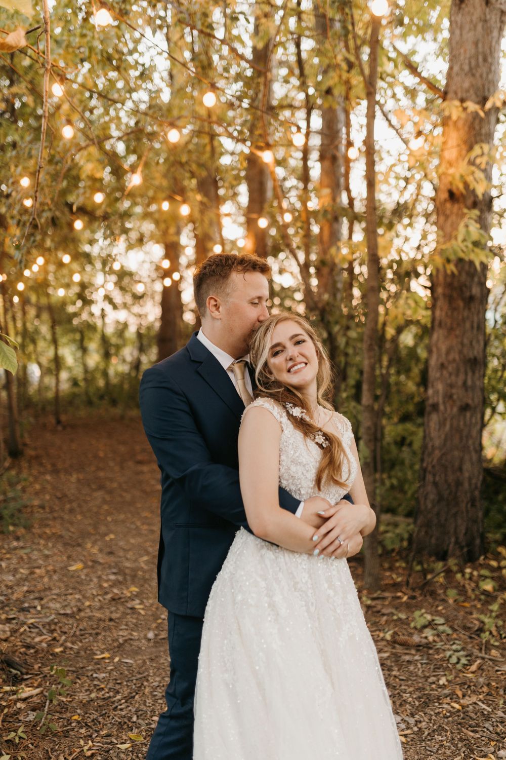 Groom kissing smiling bride on cheek under warm string lights in autumn forest.