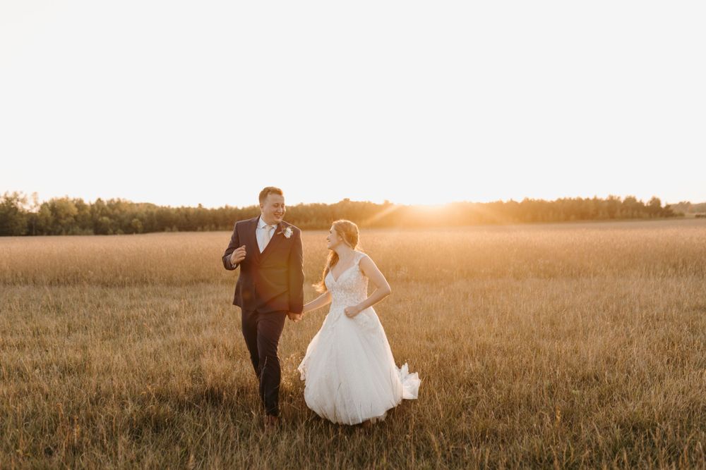 Happy bride and groom laughing while walking through golden wheat field at sunset on wedding day.