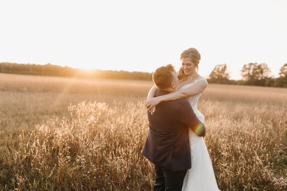 Bride smiling joyfully as groom lifts her in golden wheat field at sunset wedding photo session.