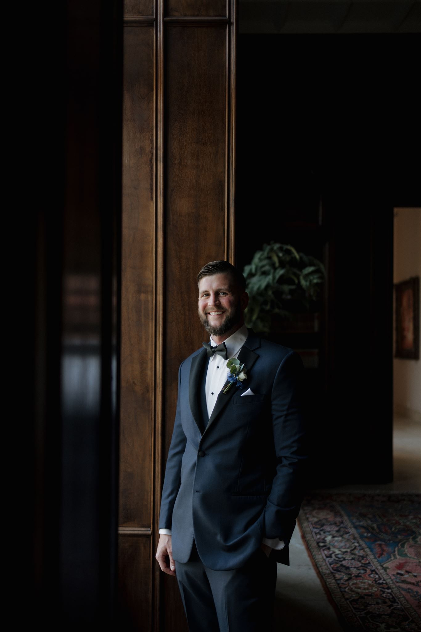 A groom in a classic navy tuxedo with bow tie stands in a dark, moody indoor setting with wooden paneling.