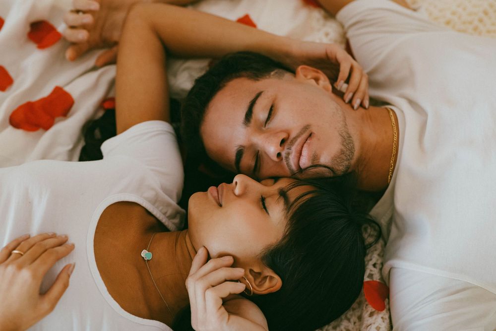 Romantic couple lying together on white bedding surrounded by red rose petals, eyes closed, smiling.