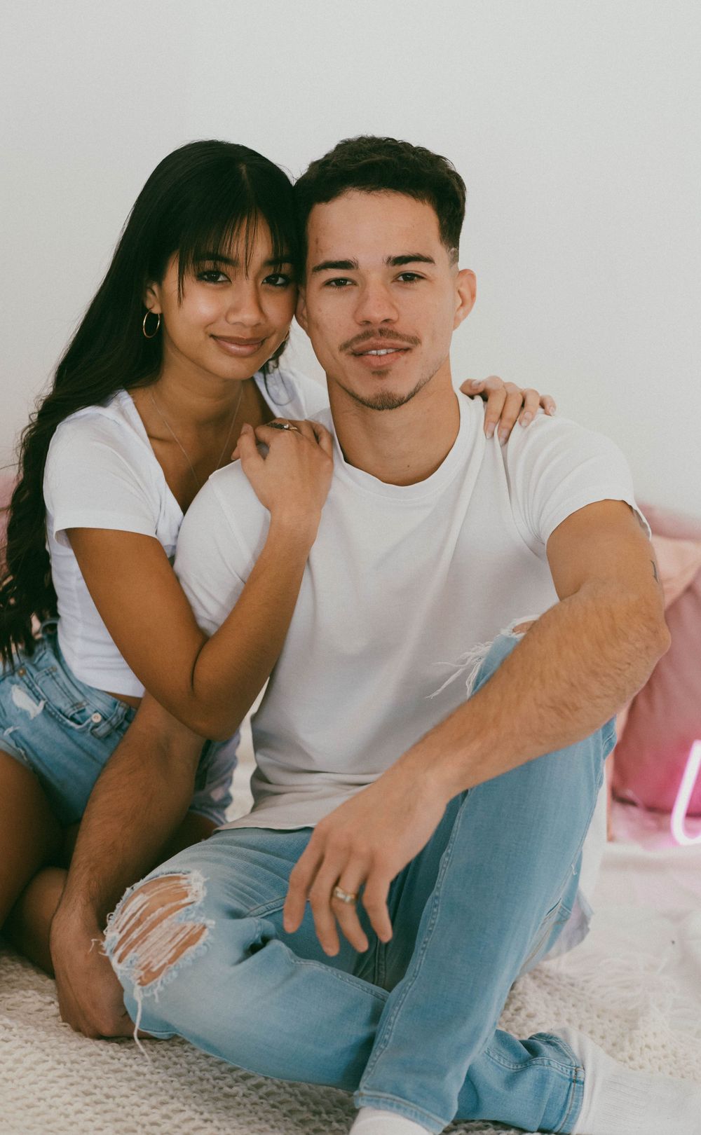 Smiling young couple in matching white tees and jeans sitting together in a cozy indoor setting.
