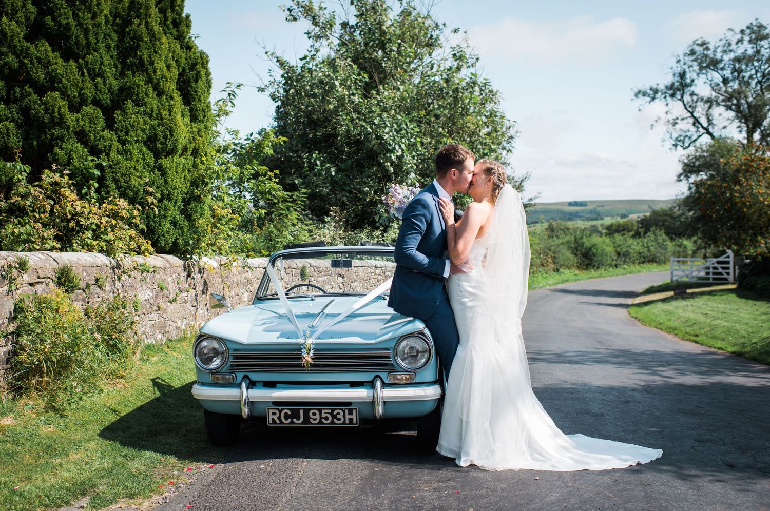Bride and groom in wedding attire sharing a kiss whilst leaning against a classic convirtable.