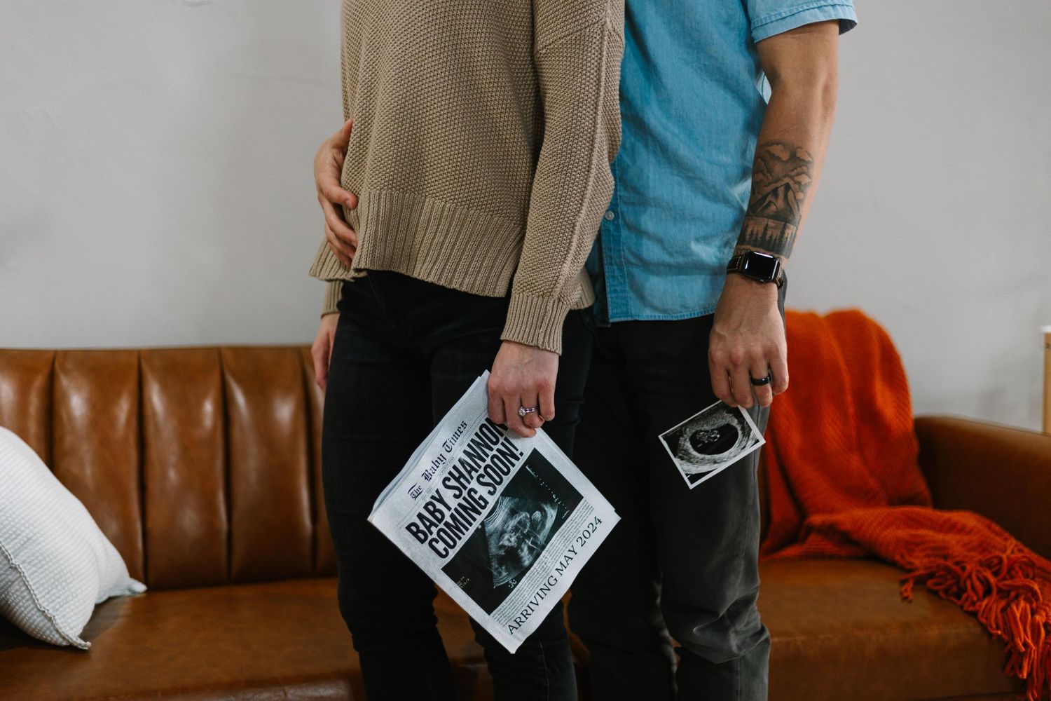 A couple holding ultrasound photos while standing near a brown leather couch.