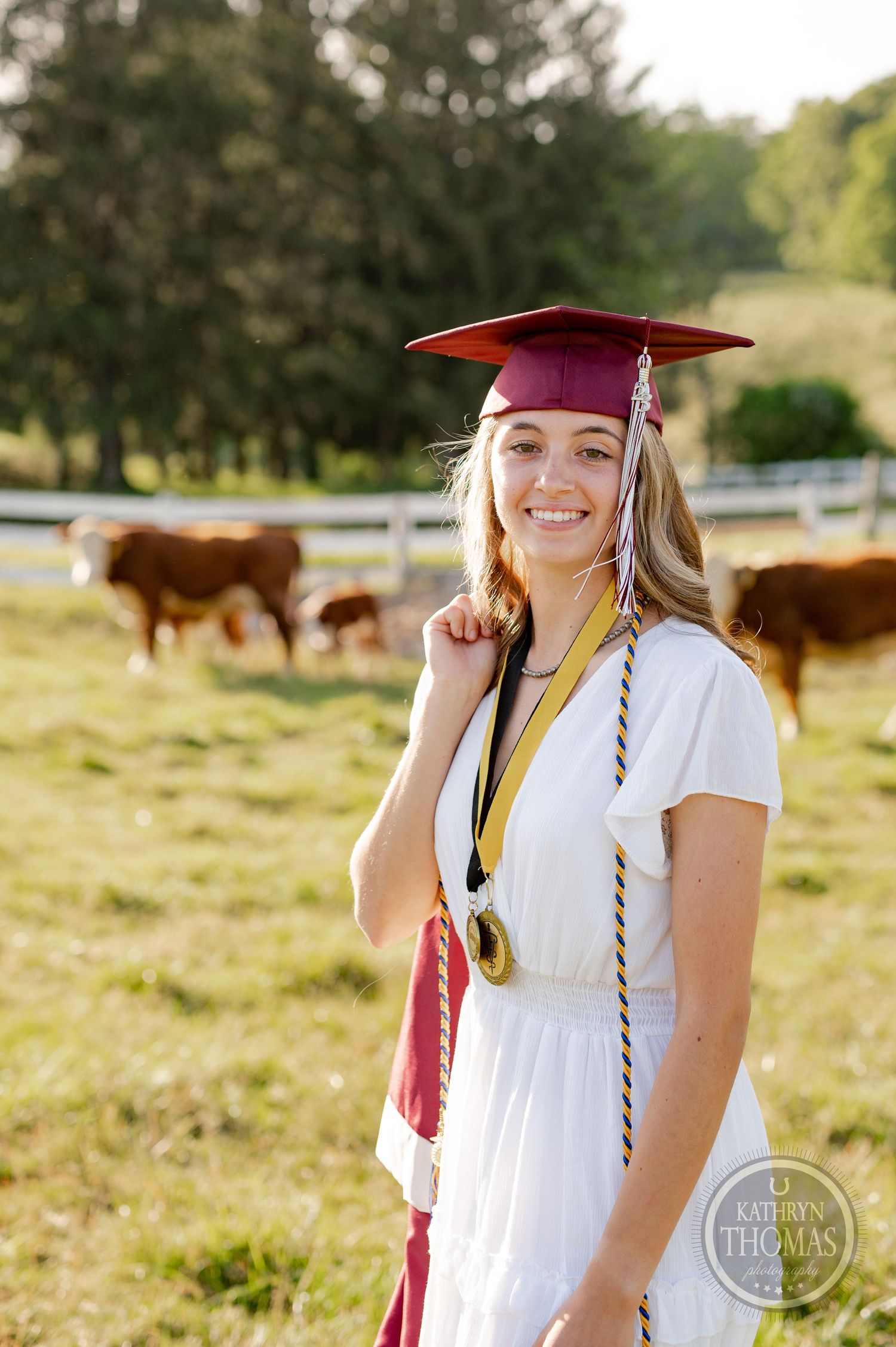 Graduate in cap and gown poses in field with grazing cattle in background during sunset.