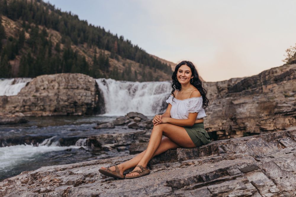 Person sitting on rocks near scenic waterfall wearing casual summer outfit.