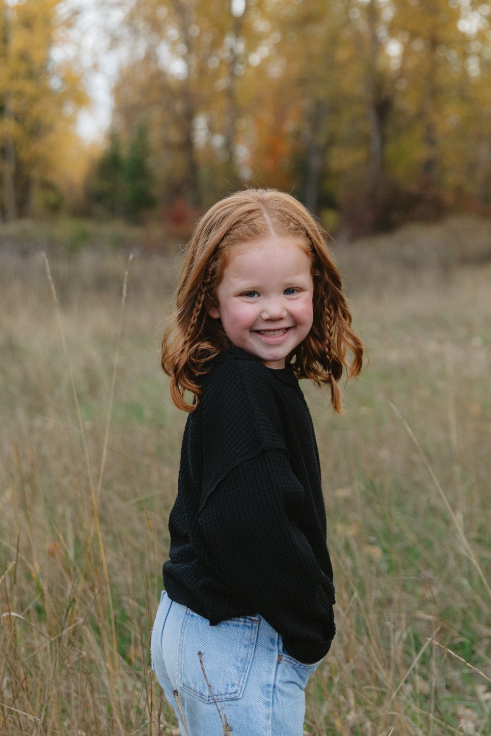 A young child in a black sweater and jeans poses in a field during autumn.