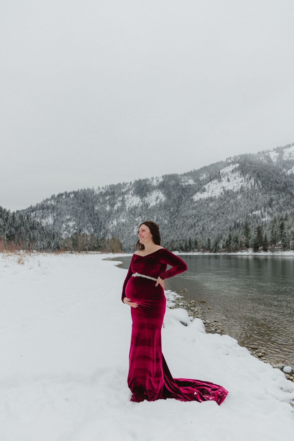 A pregnant woman in flowing red dress posing by snowy mountain lake.