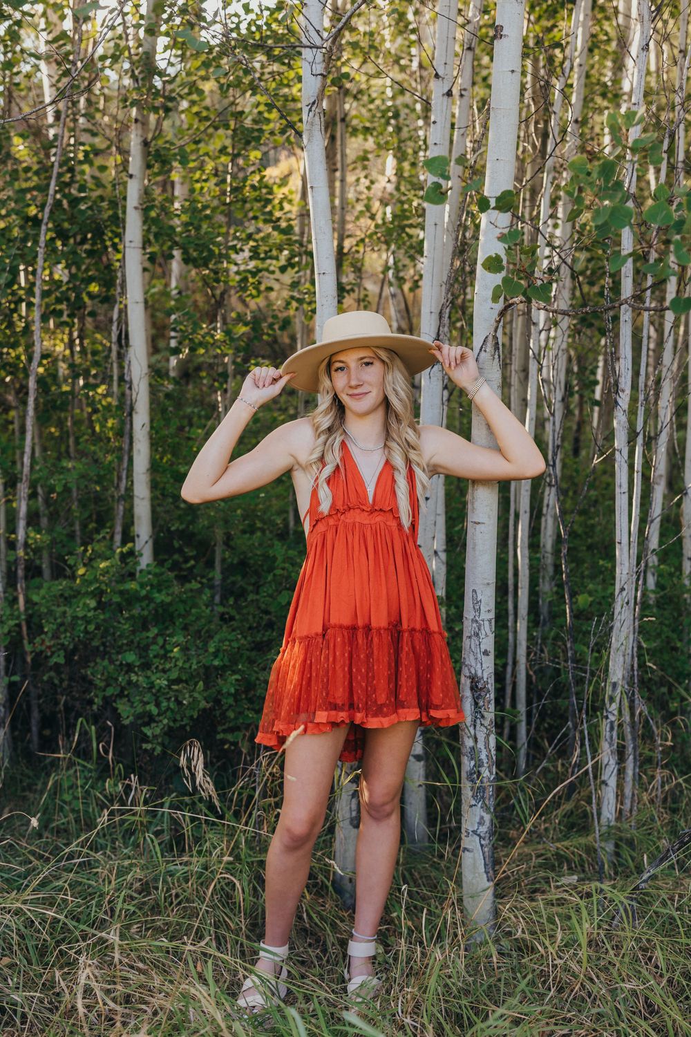 Woman in a flowy orange dress poses among aspen trees in a forest setting.
