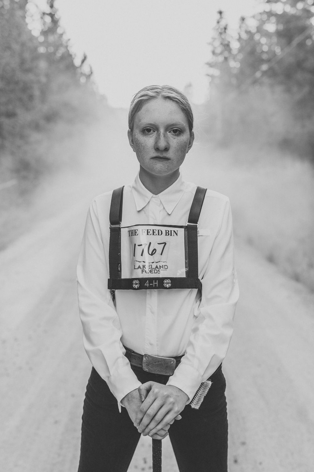 Artistic black and white portrait showing a person wearing white shirt and suspenders holding an identification number outside.