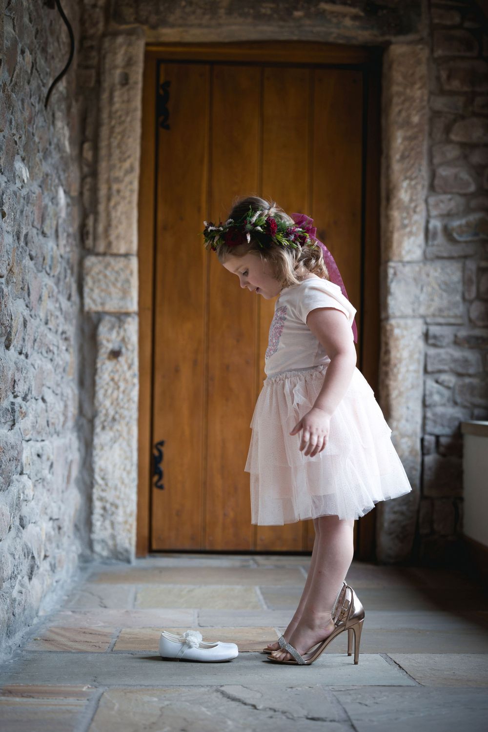 Small figure in a pink dress and floral crown looking down at shoes in a stone hallway.