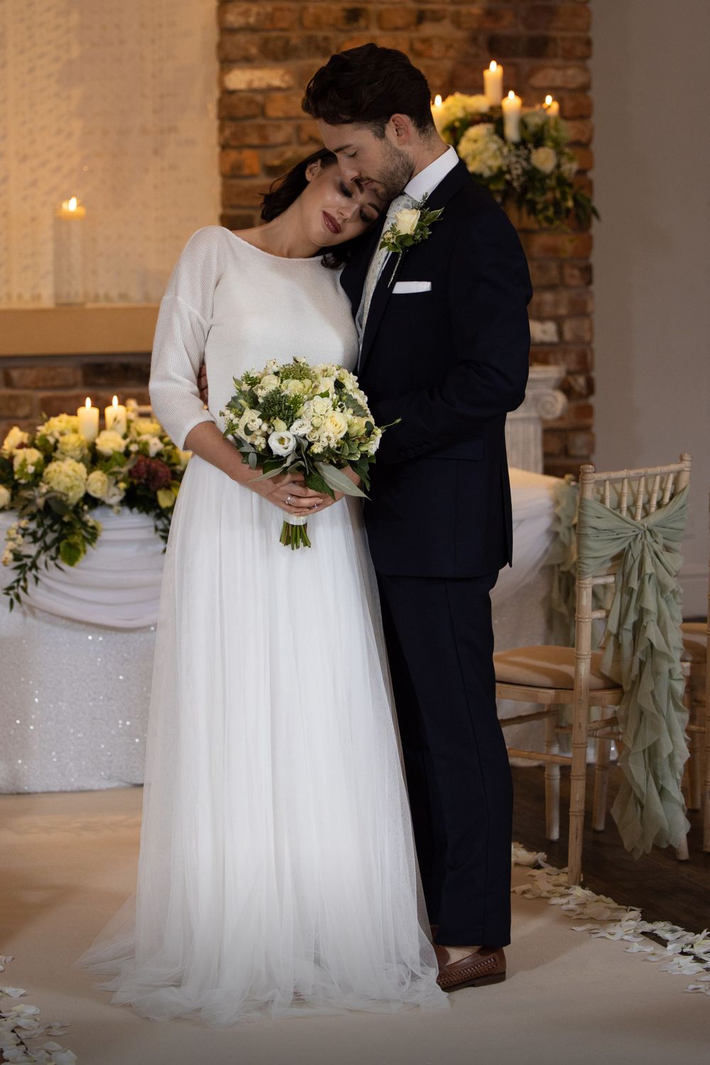 Romantic moment between newlyweds with white floral bouquet against brick wall.