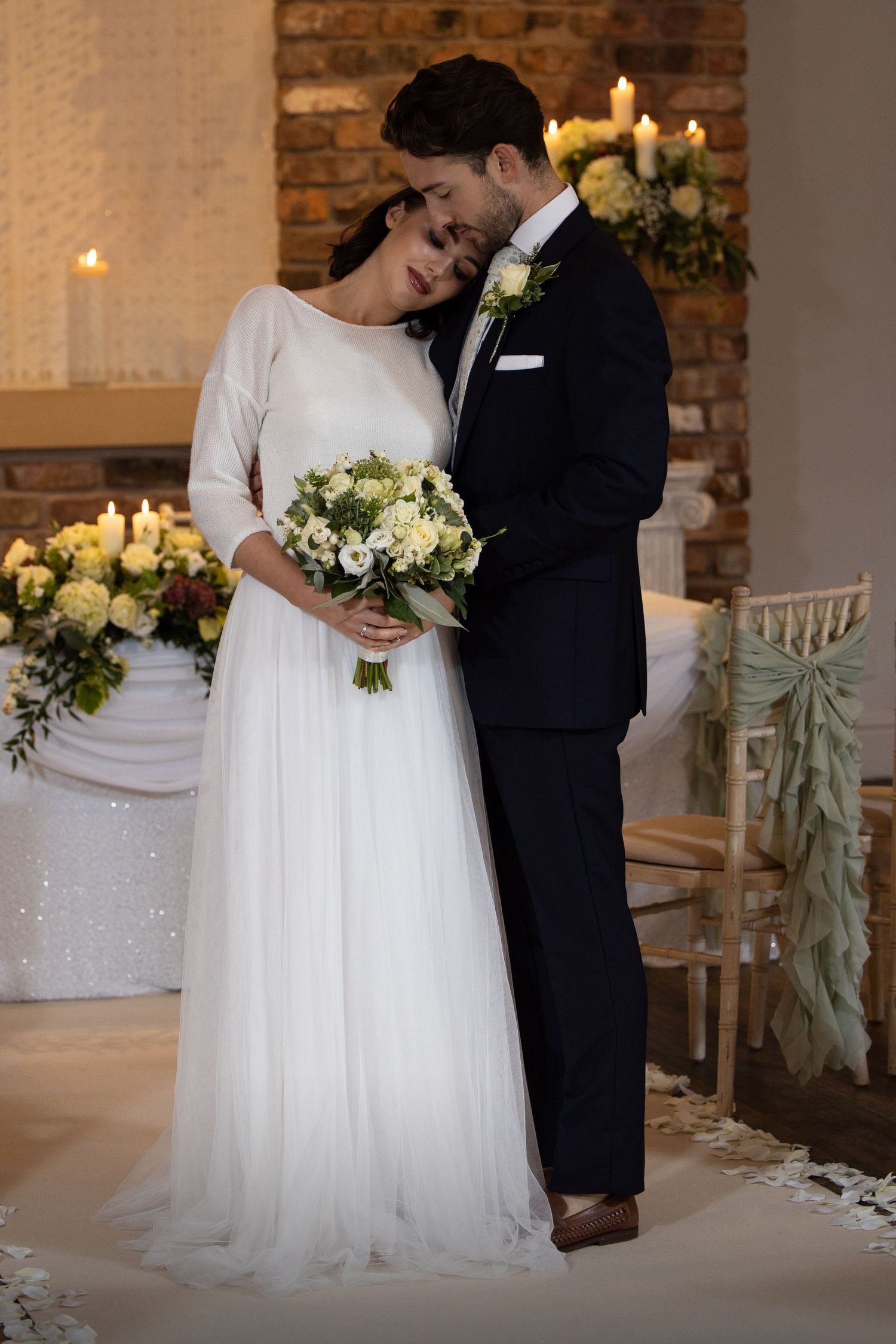 Romantic moment between newlyweds with white floral bouquet against brick wall.