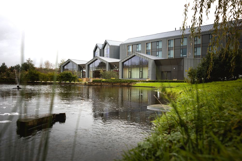 A modern hotel building reflected in a pond surrounded by landscaped grounds.