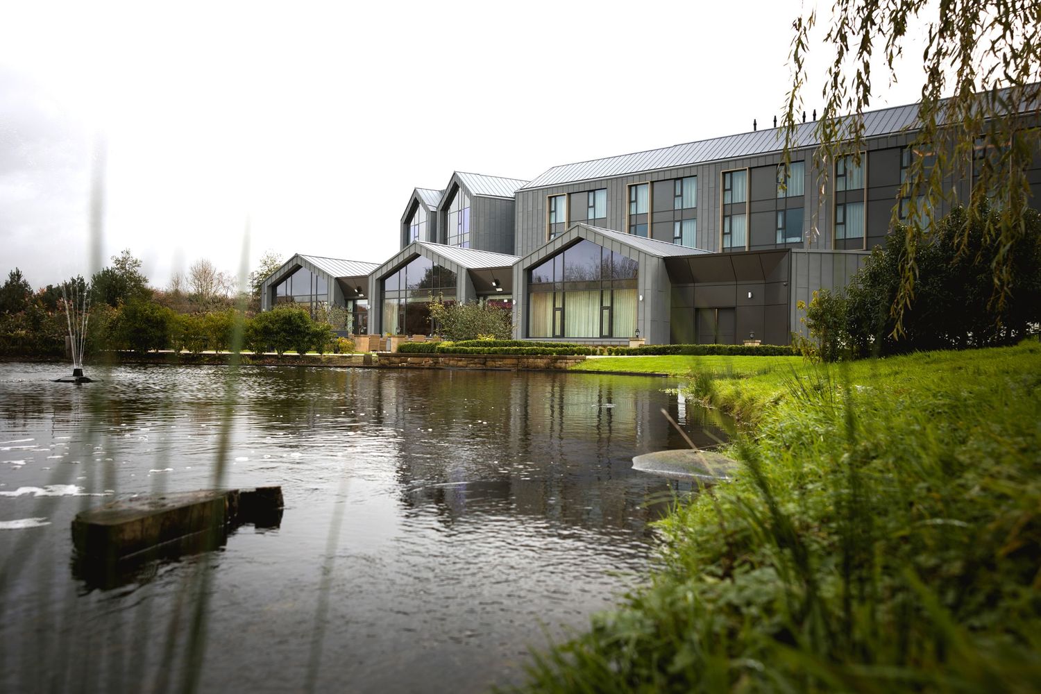 A modern hotel building reflected in a pond surrounded by landscaped grounds.