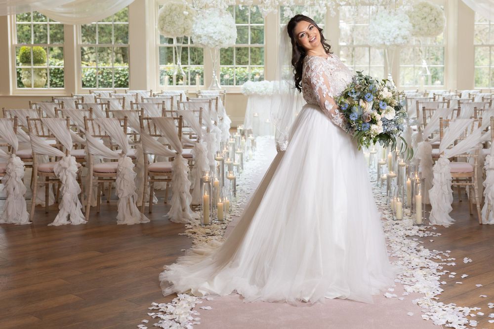 Bride posing in elegant white gown in sunlit wedding venue with decorative white chairs.