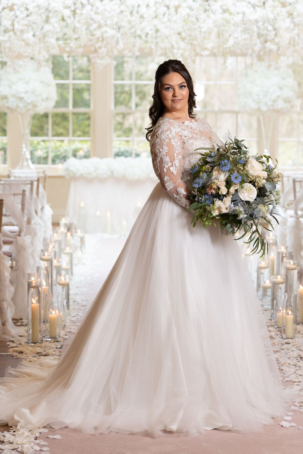 Bride in long-sleeved lace gown holding luxurious blue and white bouquet in candlelit aisle.