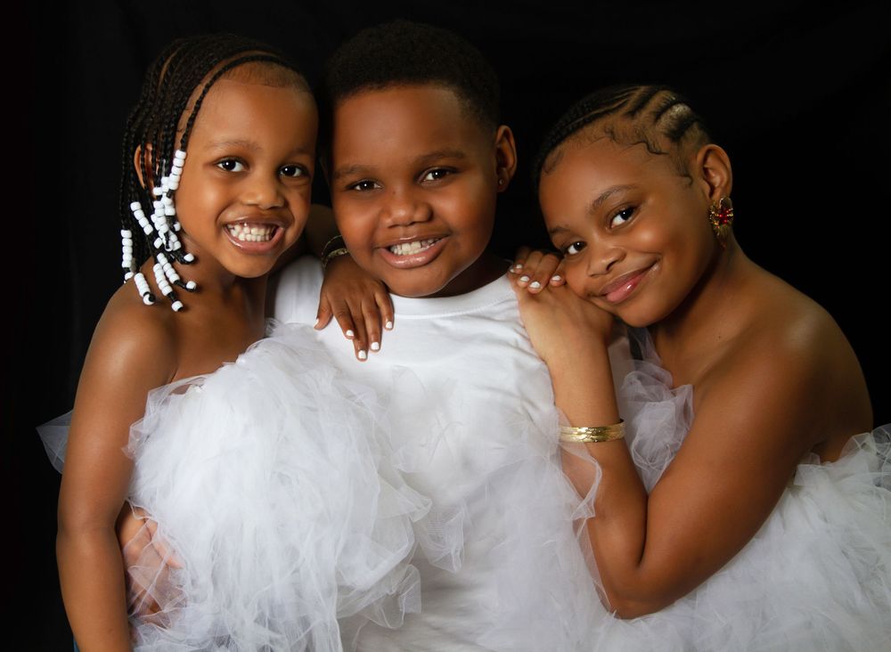 Lifestyle portrait of three children in matching white dresses against dark background.