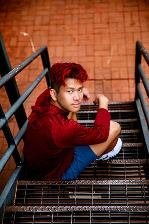 Person in red hoodie sitting on metal stairs against brick wall background.