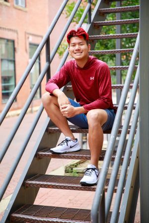 Person in red shirt and blue shorts sits casually on metal stairs with brick wall background.