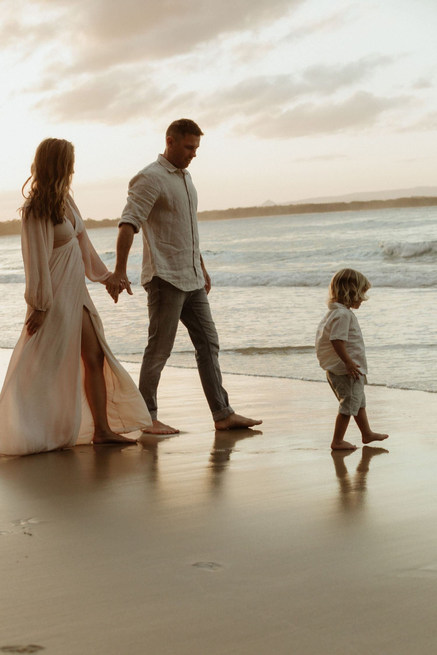 A family walks together on a beach at sunset, leaving footprints in the wet sand.