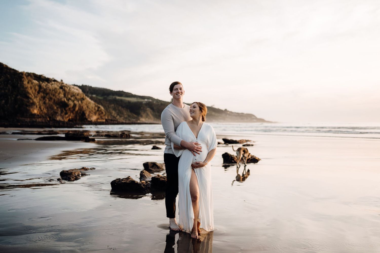 Romantic couple embracing on rocky beach at sunset with ocean waves in background.