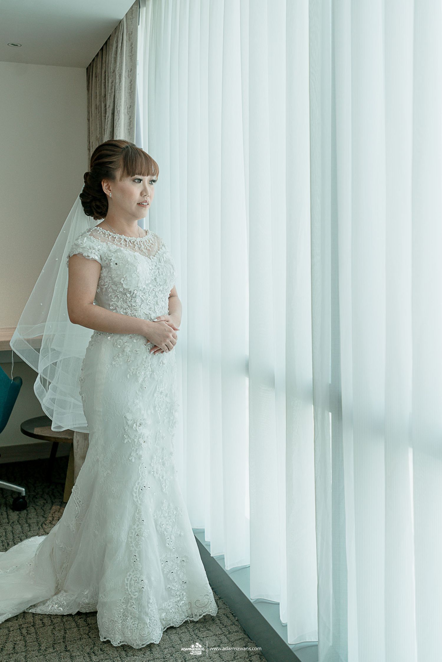 A bride in an elegant lace wedding gown stands by a bright window with flowing curtains.
