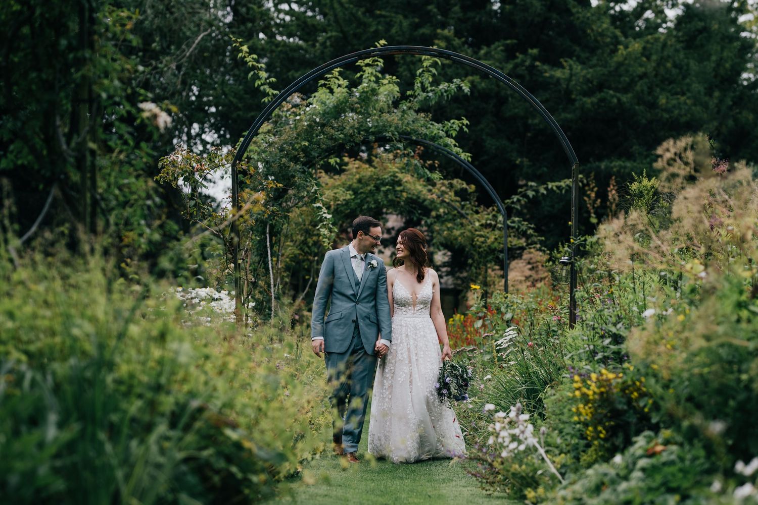 Newlyweds walk together through a lush garden pathway surrounded by flowering plants and greenery.