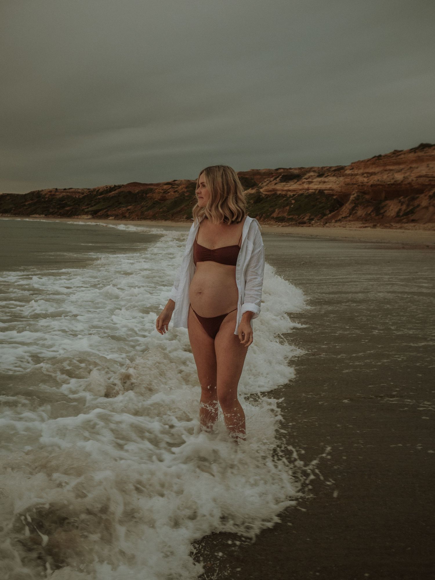 Person in white shirt and swimwear standing in ocean surf near red cliffs.