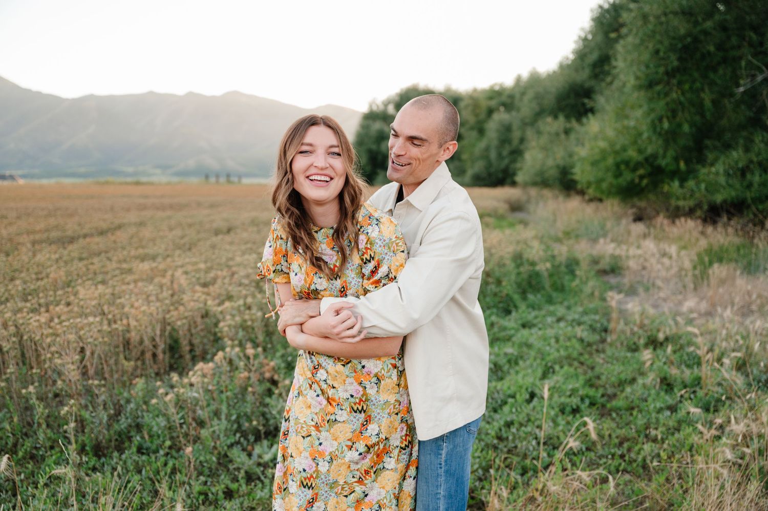 Couple embraces in a rustic field with mountains and forest in the background during golden hour.