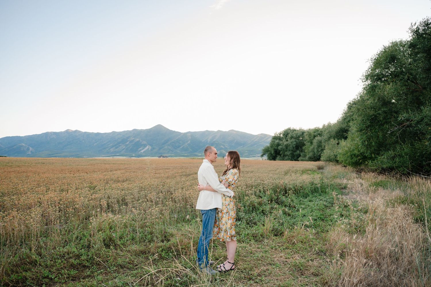 Romantic couple stands together in a golden field against a backdrop of mountains and trees.