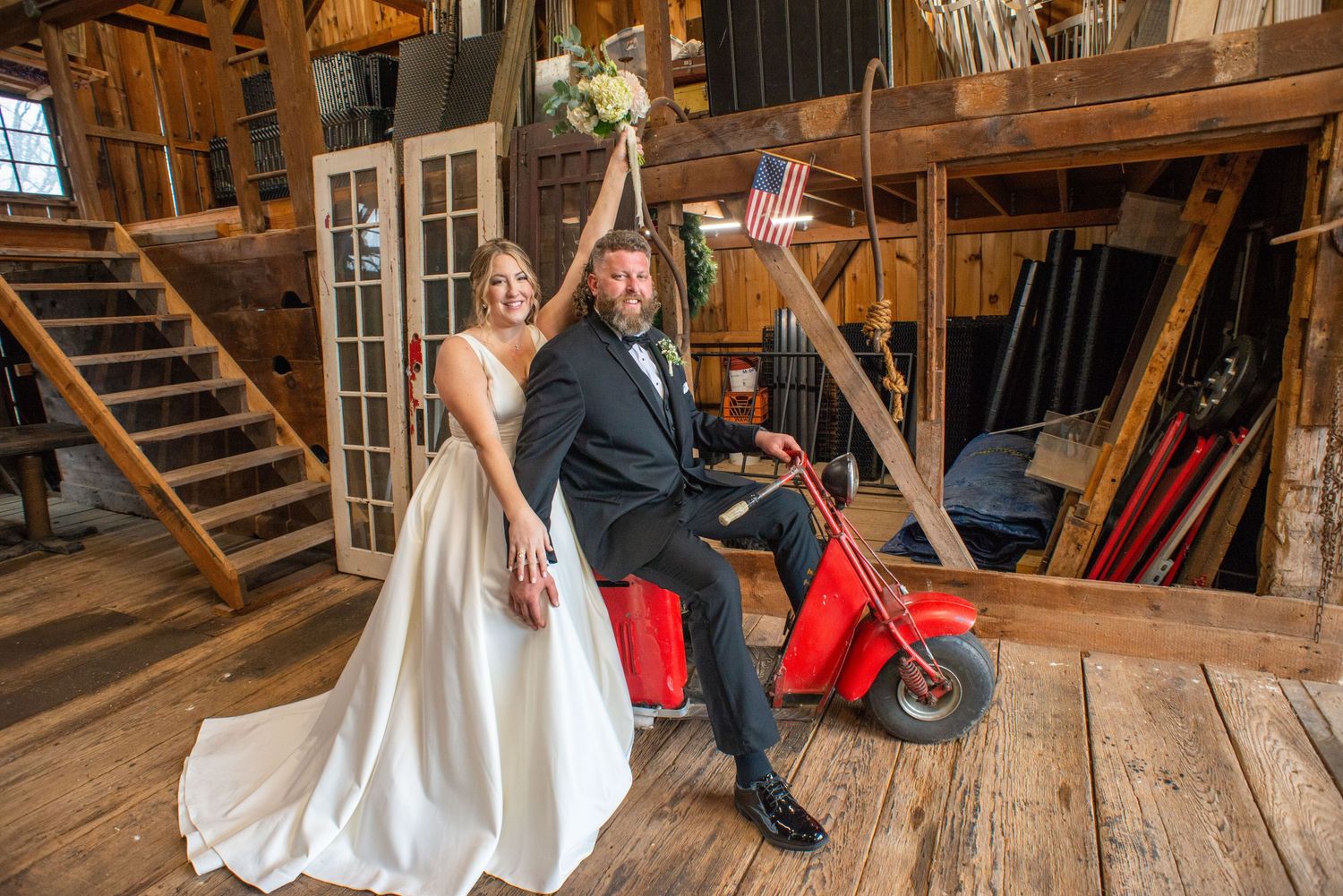 A newlywed couple poses playfully on a red Vespa scooter inside a rustic barn setting.