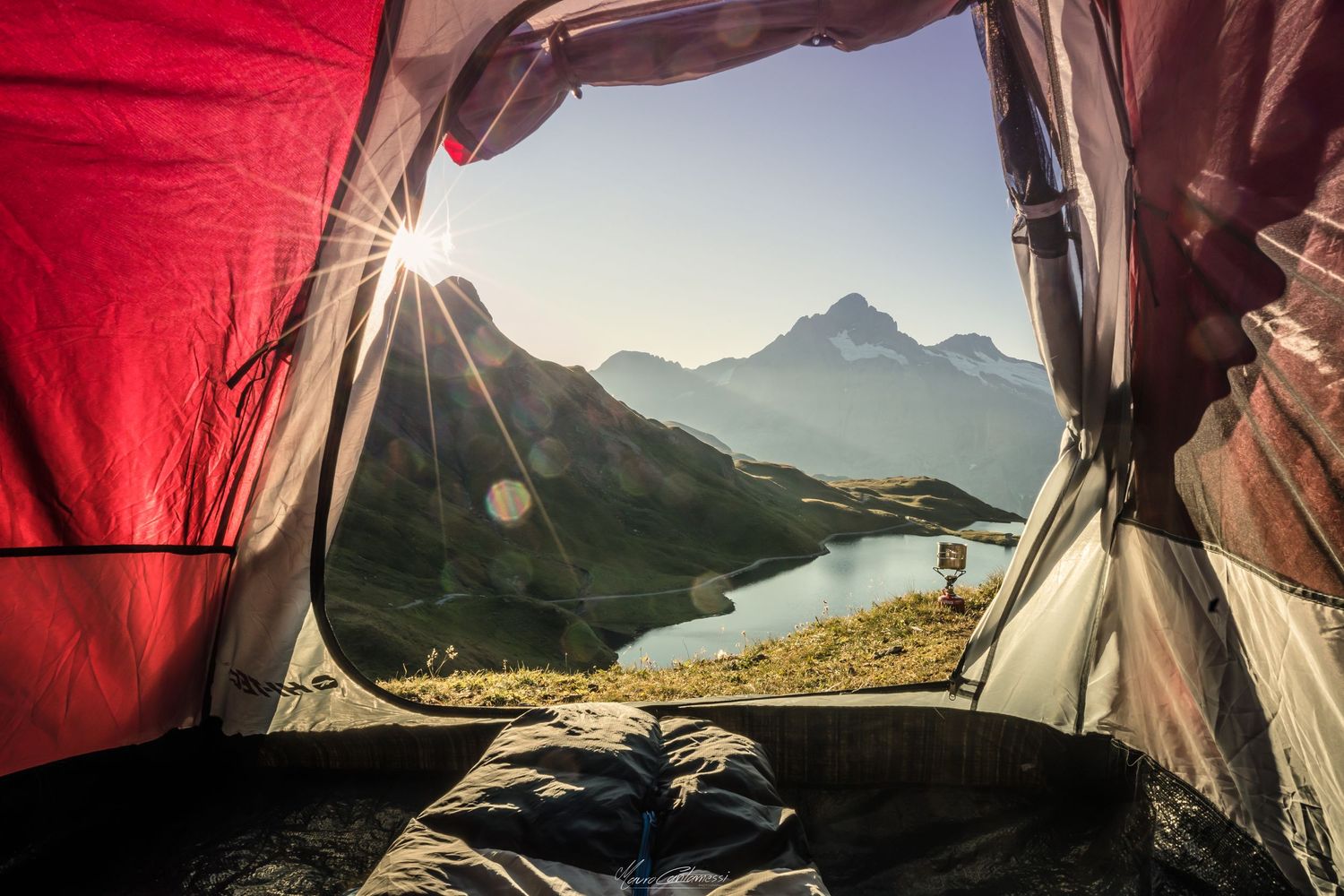 Mountain lake and peaks viewed through tent opening at sunrise with red tent flaps framing the scene.