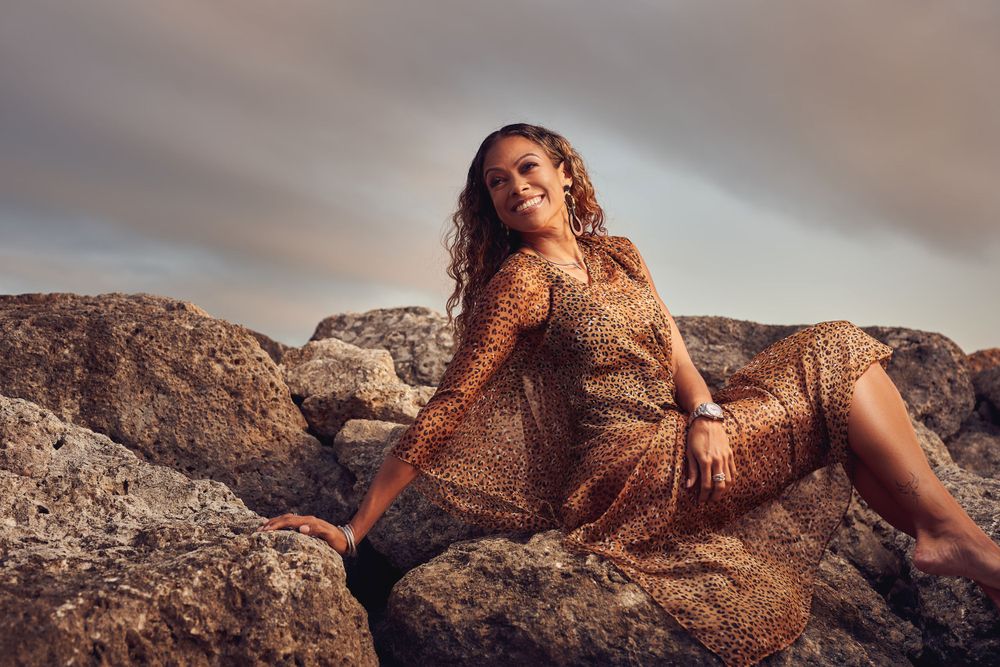 Woman in sequined gold dress poses dramatically on coastal rocks at sunset.