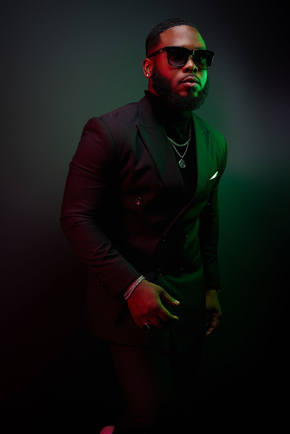Stylish figure in dark suit and sunglasses posed in dramatic lighting with red and green tones.
