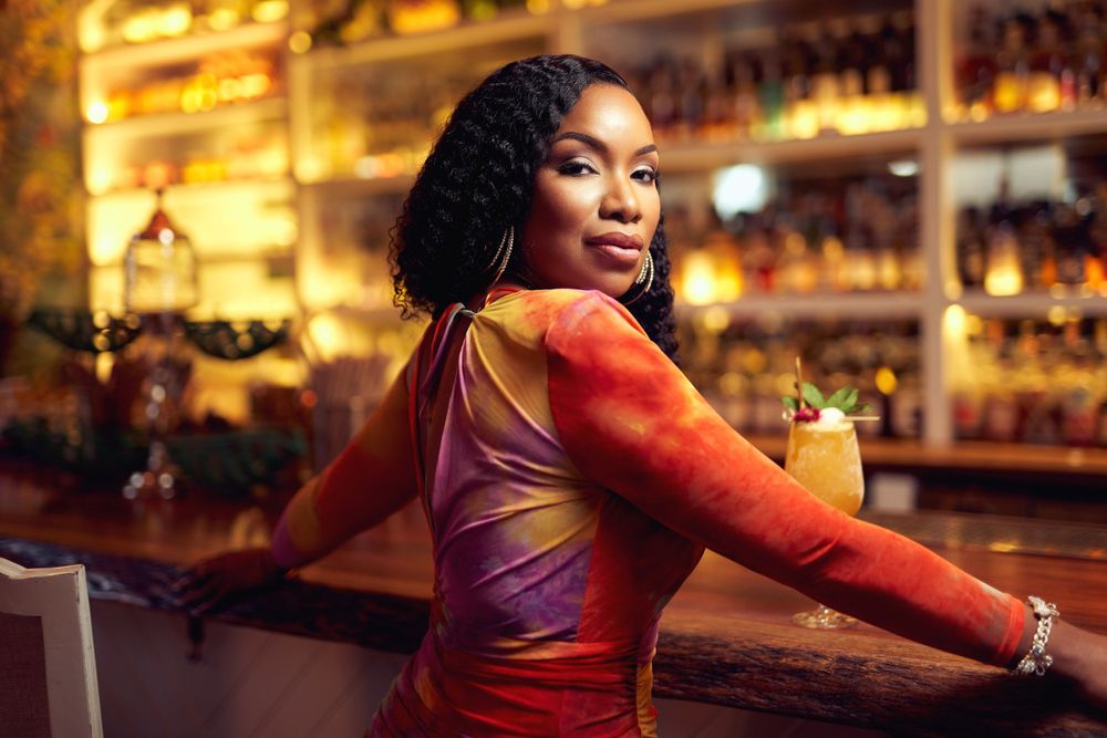 Elegant woman in red dress sitting at illuminated bar with cocktail.