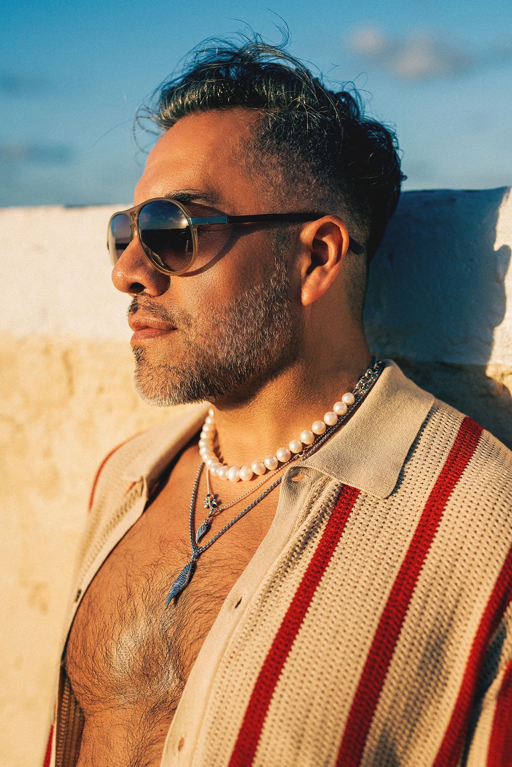 Person in striped sweater and sunglasses posing in warm sunlight against white wall.