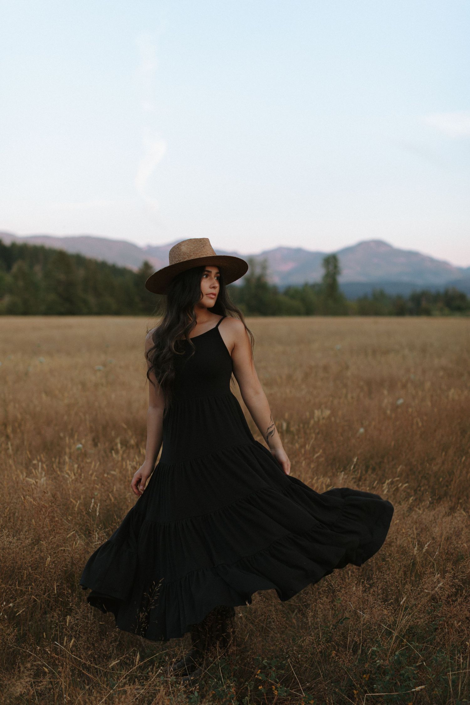 A woman in a flowing black dress twirls in a golden field with mountains in the background.