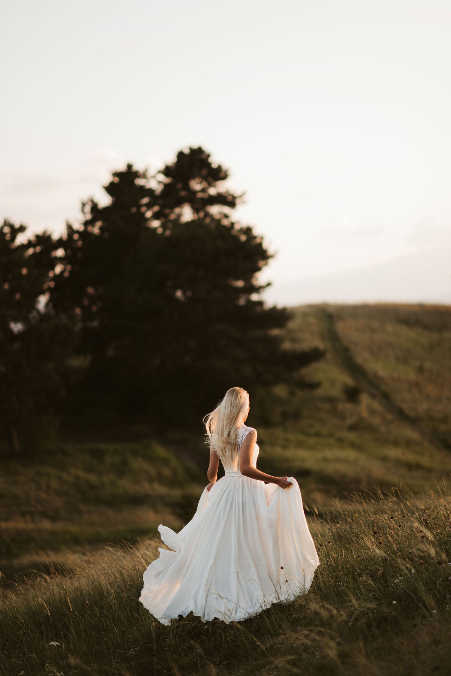 White wedding dress flowing in the wind on a grassy hilltop at sunset.